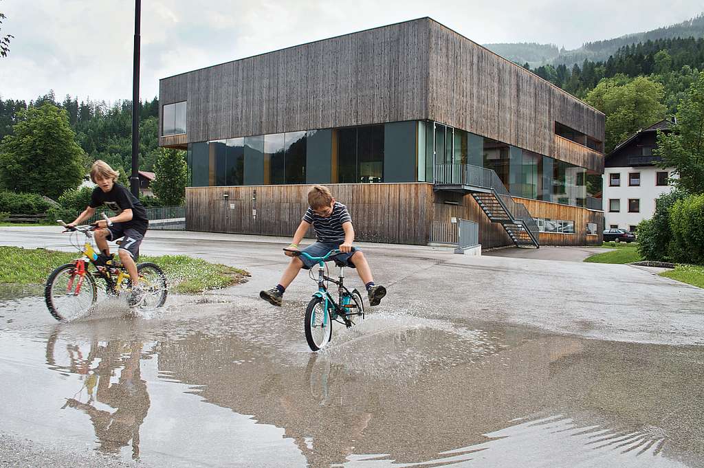 Zwei Kinder fahren vor dem Gemeindeamt Hinterstoder mit Fahrrädern durch Regenpfützen.